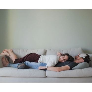 Para leżąca razem na jasnej sofie, kobieta uśmiechnięta oparta na mężczyźnie z poduszką na oczy, w tle jasna ściana i spokojna, domowa atmosfera.